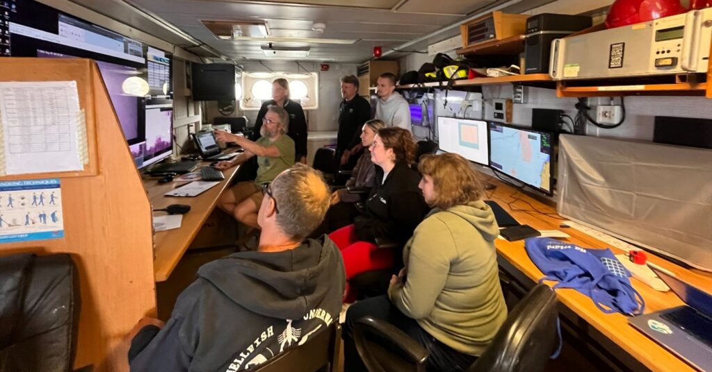 people sat down looking a video footage on screens in a room on the ship